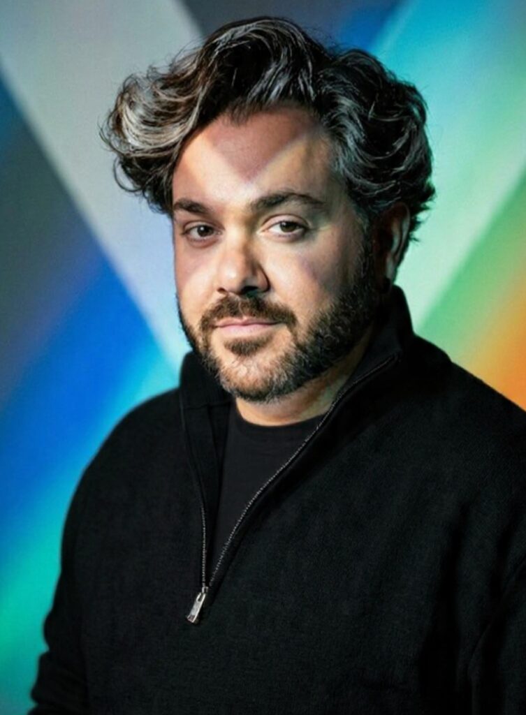 Ein Mann mit lockigem Haar und Bart schaut freundlich vor farbigem Hintergrund